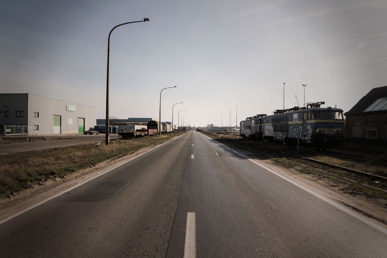 An empty road with train tracks running parallel on the right side, featuring old locomotives parked beside industrial buildings under a clear sky.