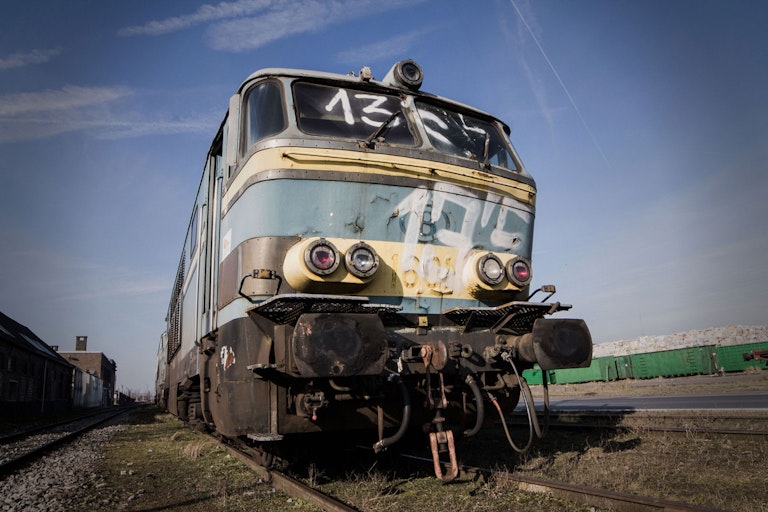 An old, worn-out locomotive with chipped blue and white paint stands on train tracks, under a clear sky. The front displays the number "43" and has multiple headlights.