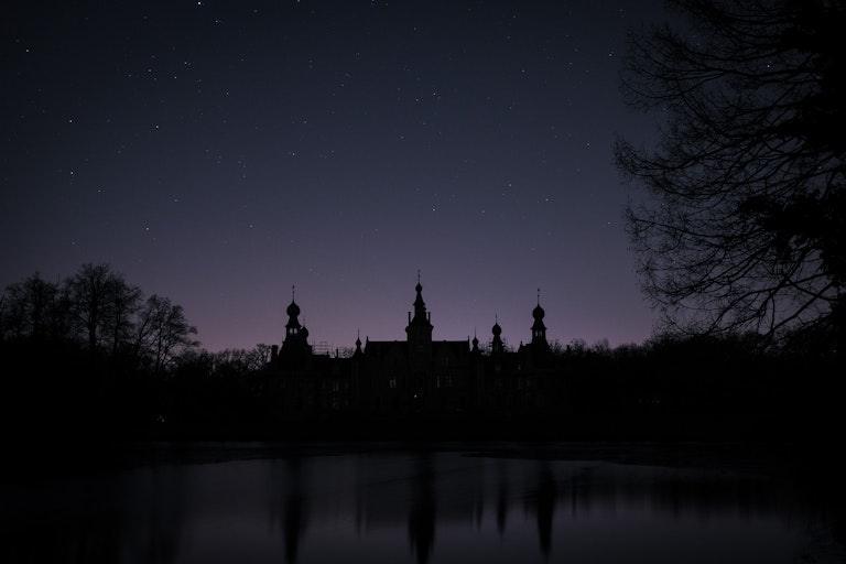 A silhouetted castle with multiple spires reflected in a still lake under a starry night sky, with trees visible on the right edge.